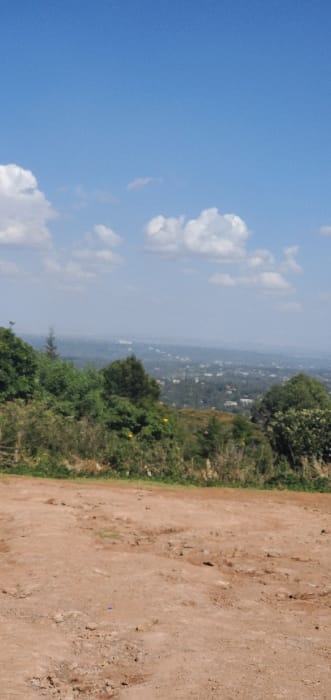 The image presents an elevated panoramic view from Ewuaso Oonkidong'i ward, Kenya. The foreground consists of an expanse of reddish-brown, dry earth, characterized by uneven surfaces, ruts, and loose soil. A dense line of green vegetation, including various bushes and trees, stretches horizontally across the midground. Beyond this foliage, a sprawling urban or semi-urban landscape is visible in the background, featuring numerous indistinct buildings and a faint linear structure possibly indicating a road or elevated thoroughfare, all partially obscured by atmospheric haze. The sky occupies the upper portion of the frame, displaying a clear, bright blue hue with scattered white cumulus clouds. No people, animals, or specific objects are present in the foreground or midground, and no actions or interactions are discernible.