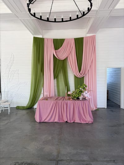 The image shows an interior decorated for an event, likely a wedding reception. A table is draped in mauve fabric, adorned with a floral arrangement of green and white flowers. Behind the table, drapery is arranged in alternating stripes of pale pink and olive green fabric. The room is a modern space with white walls and a grey concrete floor. A large chandelier hangs from the ceiling, and a botanical design is etched on the wall. The location is presumed to be in Юг, Russia. There are no people visible in the image. The scene focuses on the decorative elements and suggests a celebratory occasion.