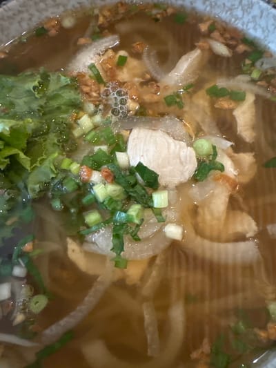 A close-up, top-down view of a bowl of pho soup reveals a savory broth filled with thinly sliced white onions, pieces of cooked chicken, and chopped green onions. Noodles are visible beneath the surface, partially submerged in the rich, brown liquid. Some fried garlic bits are scattered throughout the soup, adding texture and flavor. A portion of bright green lettuce is also present, offering a fresh contrast to the cooked ingredients. The dish is presented in a speckled gray bowl. The origin of the image is an unspecified restaurant in Siem Reap, Cambodia.