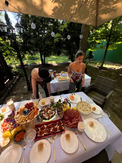 The image depicts an outdoor dining scene, possibly in a backyard, with a table laden with food and drinks. A white tablecloth covers the table, which is set with white plates, cups, and cutlery. Among the food items are platters of sliced meats, cheeses, breadsticks, olives, fruits, and what appears to be a salad. There are also small appetizers arranged on a tray. Two women are present; one is bent over the table, appearing to adjust or serve food, while the other stands and looks down at her phone. The setting is surrounded by greenery, with trees and bushes visible in the background, suggesting a natural or garden environment. Several string lights hang overhead, adding to the ambiance. The location is identified as Tomilino, Russia.
