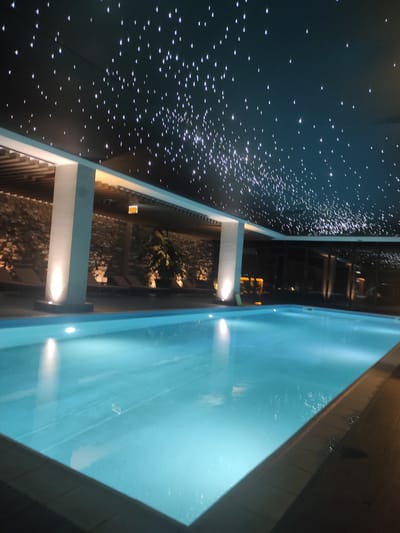 The image depicts an indoor swimming pool with a dark ceiling illuminated by numerous small, bright lights, resembling a starry sky. The pool water is a luminous turquoise color, with reflections of the lights visible on its surface. The pool area appears to be indoors, with columns supporting a covered ceiling. In the background, there are lounge chairs and a stone-textured wall. The scene is lit with ambient lighting, casting a serene glow over the pool and surrounding area. There are no people visible in the image. The location is identified as Gudauri, Georgia.