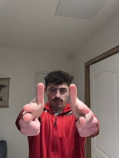 A young man with curly hair, glasses, and a mustache poses in a red jacket. He holds his hands up in a "finger gun" gesture, directed towards the camera. The setting appears to be an indoor room with white walls and a doorway visible in the background. A map is partially visible behind him. The photo has a casual, informal quality.