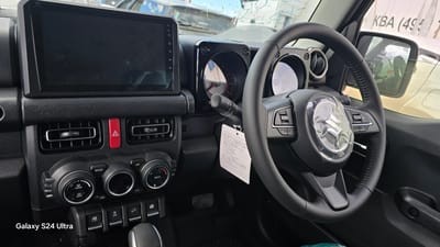 The image shows the interior of a car, likely a Suzuki Jimny, as indicated by the logo on the steering wheel. The dashboard features a central touchscreen display, climate control knobs and buttons, and an air vent. A hazard light button is also visible. The steering wheel is wrapped in protective plastic, suggesting the car is new or has recently undergone some work. A tag with Japanese writing is hanging from the steering column. The driver's side door and side mirror are also visible. Outside the car, through the windows, parts of other vehicles and a building can be seen, with some graffiti or markings on the building's exterior. The image is a close-up shot focusing on the car's interior. There is no indication of the car's location in Yakutsk, Russia, in the provided image or its crops.