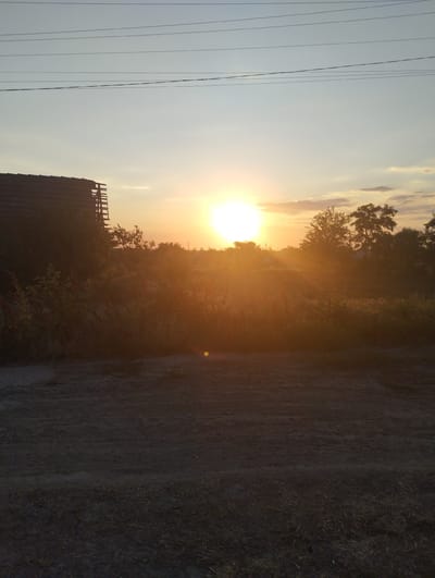 This is a photograph taken at sunset in Vasylivka, Ukraine. The scene depicts a rural landscape with a silhouetted building on the left and trees in the background. The sun is low in the sky, casting a warm, orange glow over the scene and creating lens flare. Power lines are visible against the pale sky. The foreground is a dirt path with some dry grass. The overall mood is peaceful and serene, characteristic of a sunset in the countryside. There is no visible text in the image.