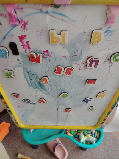 The image displays a close-up view of a child's magnetic whiteboard easel, covered in scribbled drawings and magnetic letters. The whiteboard is framed by a yellow border and is part of a larger easel structure with a teal-colored tray at the bottom, filled with chalk and other drawing supplies. The background appears to be an indoor setting, likely a home, with the floor visible at the bottom. There are no people clearly visible in the image, but the presence of children's toys and drawings suggests it is a child's play area. The scene is characterized by a chaotic yet playful arrangement of colorful letters and abstract drawings, indicating creative activity. The location is specified as Тулаўскі сельскі Савет, Belarus.