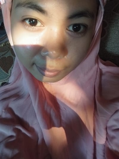 The image is a close-up, frontal, selfie-style photograph of a young woman wearing a pink hijab. The woman's face is illuminated by a harsh, diagonal shaft of light, casting a shadow across her nose and left cheek. Her eyes are open and looking towards the camera. Her lips are slightly parted and appear to be glossy. The background is a textured surface, possibly upholstery or a woven material, with a pattern of dark and light brown shapes. The woman is positioned in the center of the frame, with the light obscuring parts of her face. There is no discernible urban environment or specific location within Labissa, Indonesia, visible in the image.