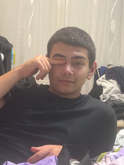 A young male with short, dark hair and dark eyebrows is shown in a medium close-up shot. He is wearing a black t-shirt and is seated, with his left arm raised and his index finger touching his right eye. His gaze is directed towards the viewer. The background consists of a light-colored curtain with vertical, wavy white patterns. To the right of the subject, a pile of folded laundry is visible, including items in white, grey, and purple. The lighting appears to be indoor ambient light. The scene is likely a residential interior, possibly a bedroom or living area. There is no discernible information to place this image specifically in Novi Iskar, Bulgaria.