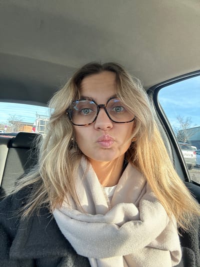 A female individual is depicted in a self-portrait taken from inside a vehicle. She has long, light blonde hair parted centrally and wears large, square, tortoiseshell-patterned eyeglasses. Her light-colored eyes are focused on the camera, and her lips are pursed in a "kissing" pose. She is dressed in a light-colored, possibly beige, scarf wrapped around her neck, over a dark-colored coat or jacket. A small gold-colored earring is visible in her right ear. The interior of the vehicle shows a light grey headliner above and a dark-colored seat behind her to the left. Through the vehicle's windows, an exterior environment is visible under a clear blue sky. Bare tree branches are present, suggesting a colder season. Several parked vehicles and what appears to be a commercial building are visible in the background, indicative of a parking lot or commercial area. The specific location within Veauche, France, cannot be definitively determined from the visual information provided in the image.
