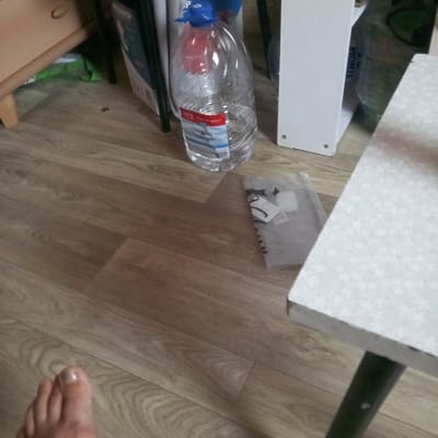The image shows a floor, a large plastic bottle of water, a small clear plastic bag with some items inside, and a white table. The floor appears to be made of wood-laminate flooring. The water bottle is partially filled with clear liquid and has a red and blue label. The clear plastic bag contains what appear to be tags or small paper items. The white table has a dark leg. The lower left corner of the image shows a person's foot with a visible toenail. The scene appears to be indoors. There is no indication of the city of Tomilino, Russia, or any other location.