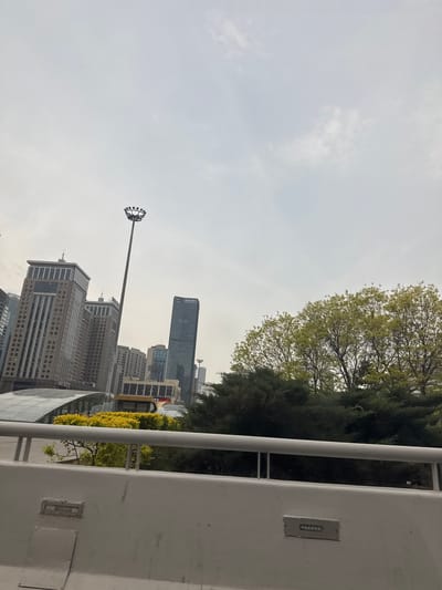 The image depicts a cityscape in the Shenhe District of China, viewed from a slightly elevated position behind a white wall or barrier. Several tall buildings of varying heights and architectural styles dominate the background. A prominent skyscraper, distinguishable by its darker facade and the visible characters on its upper section, stands out among them. In the foreground, a tall street light with multiple circular lamps extends into the sky. To the right, trees and foliage frame the urban scene. The sky is overcast.