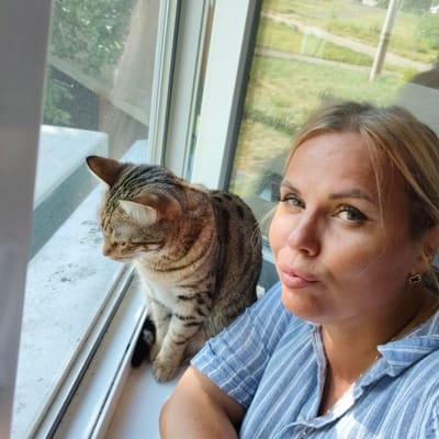A woman with blonde hair and blue eyes poses for a selfie next to a tabby cat. Both are positioned on a windowsill, with the cat looking out the window. The woman wears a blue and white striped shirt and a small, clover-shaped earring. Behind them, a screened window reveals a blurred outdoor scene with green foliage and a pathway. The environment suggests an indoor setting with a view of a suburban or semi-rural area. The image is a candid snapshot, likely taken in Dzerzhinsk, Russia, given the context.