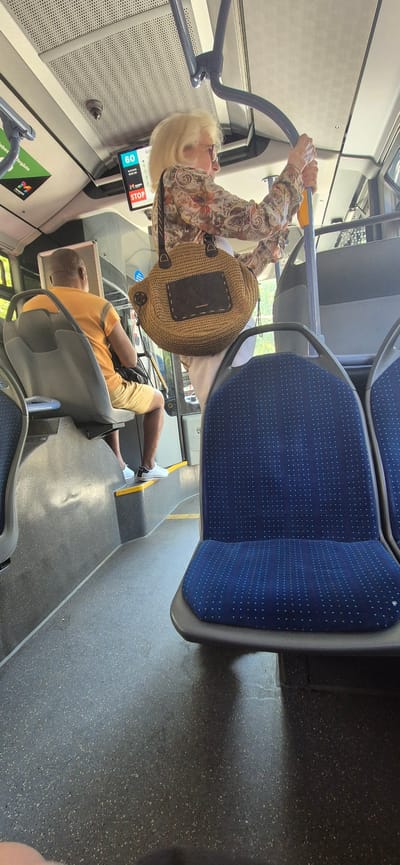 The image depicts the interior of a public transport vehicle, likely a bus, in Mainz, Germany. In the foreground, a blue seat with white speckles is visible. Behind it, a person with blonde hair and glasses, wearing a patterned shirt and holding a straw bag, stands near a pole and what appears to be a digital display showing "60". To the left, a person wearing an orange t-shirt and yellow shorts is seated. The floor is dark and speckled, and the ceiling has a textured appearance. A sign with the "M" logo and the text "Mainzer Verkehrsgesellschaft" is visible on the left side, indicating the location is within Mainz.