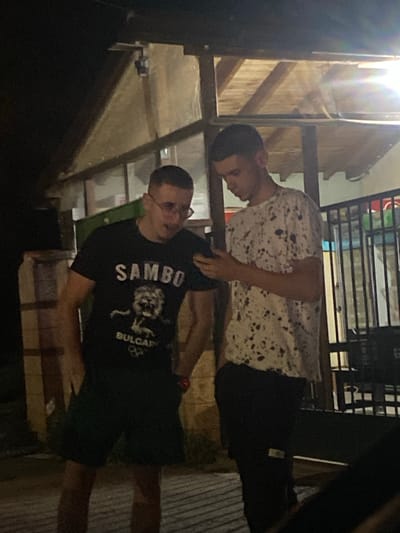 Two young men stand outdoors at night. The man on the left wears a black t-shirt with the word "SAMBO" and a graphic of a lion above "BULGARIA". He also wears dark shorts. He is looking down at a mobile phone he is holding. The man on the right wears a white t-shirt with a black splatter pattern and dark pants. He is also looking down at the phone. They appear to be standing near a building with a wooden overhang. The location is Gorna Malina, Bulgaria.