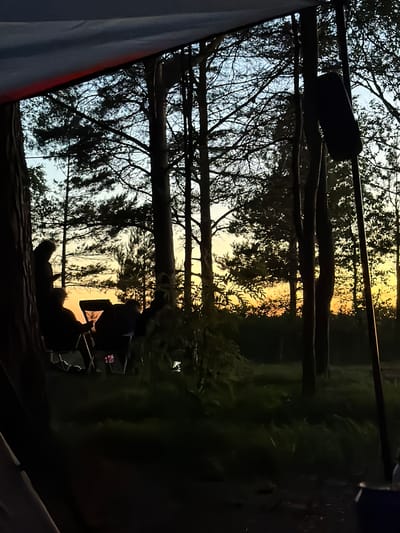 The image depicts a campsite at dusk in Meremõisa, Estonia. Silhouetted figures are seated in camping chairs, suggesting a group gathering. The scene is characterized by tall trees and the soft glow of the setting sun filtering through the foliage. The foreground is dark, indicating it is within a tent or shelter, with a hint of camping equipment visible. The overall atmosphere is one of serene outdoor recreation as day transitions to night.