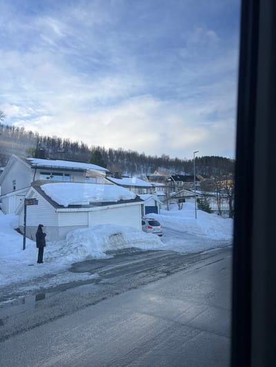 A residential street scene in Tromsø, Norway, is depicted during daylight hours under winter conditions. The foreground features a wet, partially cleared asphalt road with visible tire marks and puddles, flanked by large, irregular mounds of ploughed snow. On the left side of the road, a lone individual wearing a dark winter coat and dark pants stands beside a bus stop pole. This person faces slightly right, holding a small object in their hands, positioned next to a single-story white structure with a heavily snow-laden roof. This structure adjoins a larger two-story white house, also covered in snow. A light-colored SUV is parked in a snow-lined driveway connected to the property. In the midground, a cluster of additional snow-covered residential buildings is visible, interspersed with bare deciduous trees and a street light. The background is dominated by a steep, snow-covered hillside densely forested with trees, under a blue sky with scattered white clouds.