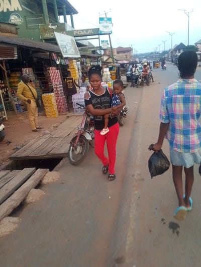 The image depicts a street scene in Orlu, Nigeria. A woman in a black shirt and red pants carries a child in a blue dress. A person wearing a blue plaid shirt walks in the opposite direction, carrying a black plastic bag. Behind the woman, a red scooter is parked, and several people are gathered. Stacks of bottled drinks are visible next to a storefront. A man in a yellow suit stands to the left of the frame, talking on a mobile phone. The street is paved, with a wooden structure covering a drainage ditch. In the background, cars, motorcycles, and auto-rickshaws are on the road. A sign displaying "Mouka Foam" is visible above the storefront.