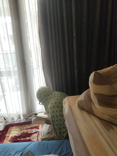 A person wearing a green headscarf with a polka dot pattern and glasses is reading a book. They are seated on a maroon prayer mat with an intricate design, which is placed on a blue surface. Behind the person, sheer white curtains are visible, suggesting a window with natural light coming through. To the right of the frame, a bed with pillows that have brown and beige stripes is partially visible. The overall setting appears to be indoors, possibly a bedroom. The natural light filtering through the curtains indicates it is daytime. The atmosphere seems peaceful and focused, with the individual engrossed in their reading. The location context is Bekasi, Indonesia. No visible text is present in the image.