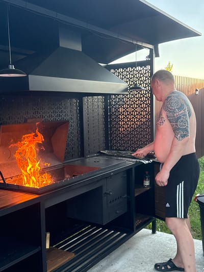 In Starokorsunskaya, Russia, a man with extensive tribal tattoos on his arm and shoulder stands beside an outdoor barbecue. He is wearing black Adidas shorts with white stripes and black sandals. The barbecue is large and black, featuring a prominent hood and a fiery grill. The man appears to be attending to the grill, holding a utensil in his right hand. Behind him, a wooden fence and some greenery are visible, suggesting a backyard setting. The sky is clear with daylight.