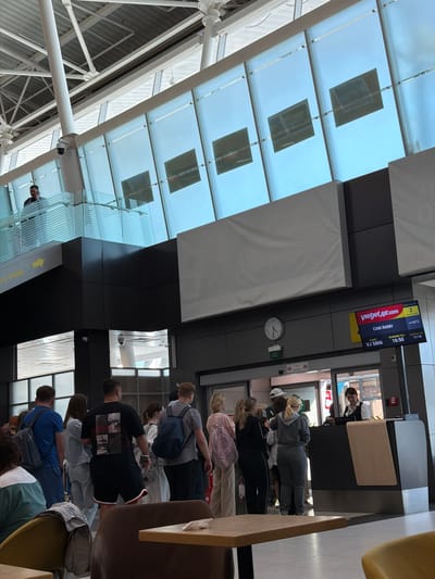 At a boarding gate in an airport in Togashevo, Russia, several passengers are seen queuing, presumably awaiting their flight. The scene is captured from a slightly elevated perspective, looking down the line of people towards the boarding counter.

In the foreground, a queue of passengers stands, many with backpacks, facing a boarding counter. An airport staff member, wearing a white shirt and black waistcoat, is visible behind the counter, looking towards the queue. To the right of the counter, a flight information display screen shows details for a Vietjet Air flight. The flight, VJ 5806, is destined for Cam Ranh, Vietnam. Boarding is scheduled until 16:50, with a departure time of 17:10. The screen also indicates a temperature of +32°C. A round clock on the wall beneath the screen shows the current time to be approximately 16:25, corroborating the late afternoon setting. A green "EXIT" sign is also visible near the clock.

The airport features modern architecture with a high, slanted glass ceiling supported by white metal beams, allowing natural light into the terminal. Reflections of the internal structure and potentially external elements are visible on the glass. There are also large, white, cushion-like panels or structures suspended from the ceiling. A yellow arrow sign, partially visible, points to the right with the text "TY FREE" and "109" also indicated from OCR, suggesting proximity to a duty-free area. The atmosphere appears orderly as passengers wait.