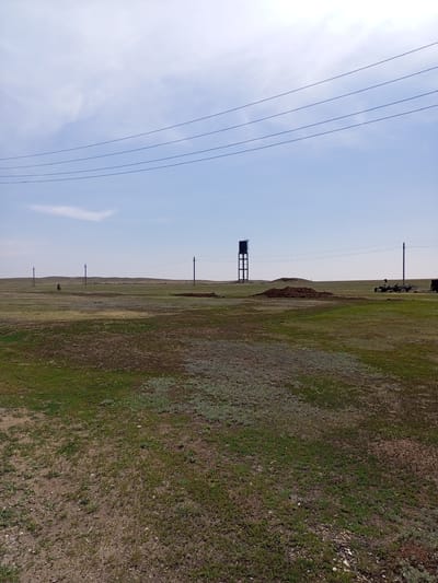 The image depicts an open, grassy field under a bright, slightly cloudy sky. In the background, a tall, dark water tower stands on a metal structure. Several utility poles with wires are visible across the landscape. A small mound of dark earth is situated in the middle ground, and agricultural machinery is present to the right. A lone figure on a bicycle is seen in the distance on the left. The terrain is predominantly flat with rolling hills in the far distance. The scene appears to be in or around the village of Ақжар, Kazakhstan, suggesting a rural or semi-rural environment.