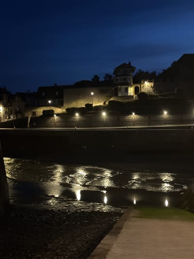 The image depicts a nighttime scene in Saint-Valery-en-Caux, France, likely along its harbor or a waterfront area. A dark, deep blue sky with subtle cloud formations forms the upper portion of the frame. Below the sky, a series of buildings are visible on a raised embankment. These structures appear to be old, with some featuring illuminated windows and external lighting. One building stands out with a distinct architectural style, possibly a house or a small inn, with multiple stories and visible light emanating from within.
A prominent stone wall or embankment runs horizontally across the mid-section of the image, separating the elevated buildings from the lower waterfront. This wall is intermittently lit by several evenly spaced lampposts, casting pools of light on the textured stone surface and surrounding shrubbery.
Below the embankment, a body of water, likely the sea or a river, is visible. The water's surface is dark and reflective, mirroring the lights from the embankment above, creating shimmering, elongated reflections. The water appears somewhat shallow in areas, revealing a dark, possibly rocky or muddy bottom in the foreground.
The foreground of the image shows a paved walkway or quay, transitioning from a darker, rougher textured area on the left to a smoother, lighter colored paved surface on the right. The overall atmosphere is quiet and serene, characteristic of a coastal town at night. No people are visible in the frame. The scene is illuminated by artificial light sources, highlighting the architectural details and the interplay of light and shadow on the water.