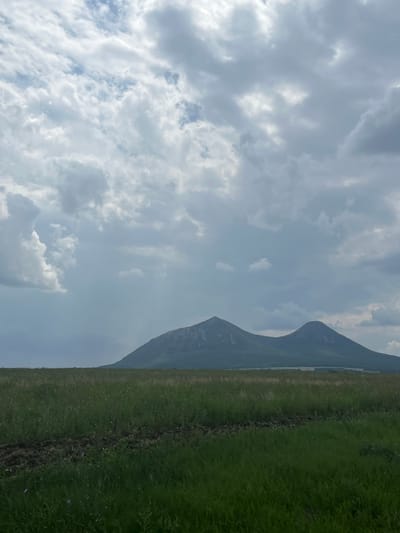 The image shows a landscape scene with two prominent, rounded mountains in the background. The sky is filled with dramatic, overcast clouds in varying shades of gray and white. In the foreground, there is a field of tall green grass with some darker soil patches visible, suggesting it might be cultivated or recently disturbed. A faint body of water is visible in the distance between the field and the mountains. The scene appears to be a natural, rural environment, potentially in or near the village of Dunaevka, Russia. There are no people or animals visible in the image.
