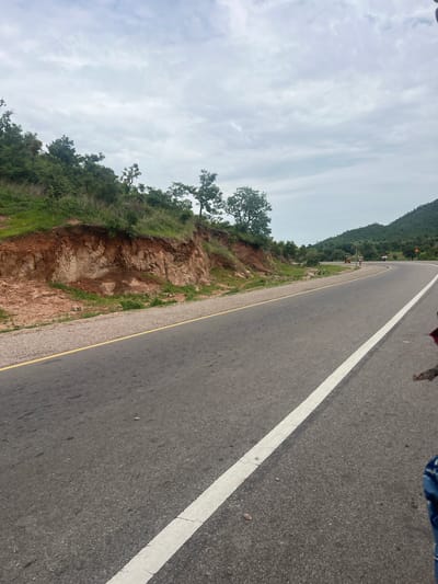 The image shows a road curving to the right, with a steep embankment on the left. The embankment is composed of reddish-brown soil and rocks, with green vegetation growing on top and along its base. The road is asphalt, with a yellow line marking the shoulder on the left and a white line on the right lane. In the distance, the road passes by rolling green hills, some of which are covered in trees. There are a few small buildings visible in the distance, as well as some signs of human activity, including a yellow vehicle and two people on the side of the road. The sky is overcast with a gray, cloudy sky. The scene depicts a rural or semi-rural road in Minda, Nigeria.