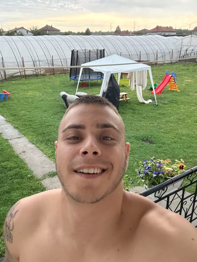 The image captures a smiling young man in a selfie-style shot, shirtless, in what appears to be a backyard or garden setting. He has short hair, light stubble, and a tattoo resembling a dragon on his left shoulder. His face, with a wide smile, dominates the right side of the frame, conveying a cheerful emotion.
The background features a vibrant green lawn traversed by a concrete pathway. Various recreational and garden items are scattered across the yard. To the left, a white canopy or tent covers a partially visible blue trampoline and possibly a covered grill. A small red and blue plastic slide and a red watering can add splashes of color further back. Near the foreground, adjacent to the man, a black metal railing is adorned with a hanging basket filled with lush purple petunias and some orange pansies.
In the mid-ground and distance, long, white, tunnel-like structures, likely greenhouses or polytunnels, stretch across the landscape, hinting at an agricultural environment. Beyond these structures, residential buildings and electrical power lines are visible under a soft, hazy sky. The horizon glows with a warm, golden hue, suggesting it is late afternoon or early evening. The setting appears to be a relaxed, semi-rural area, consistent with a location such as Slivo Pole, Bulgaria. No visible text is present in the image.