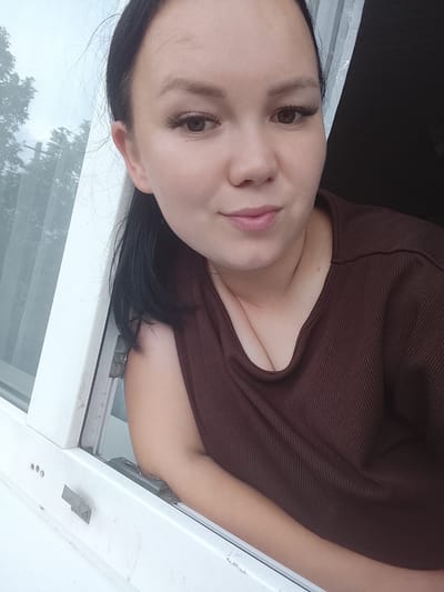 A woman with dark hair and false eyelashes is leaning out of a white window frame, looking at the camera. She is wearing a brown, ribbed, V-neck top. Her right arm is resting on the windowsill. Outside the window, trees are visible through a translucent curtain or blind. The background on the right is dark. The scene appears to be in Kuyeda, Russia, based on contextual information not present in the image itself.