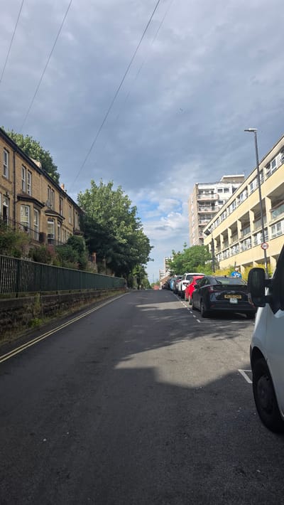 This is a medium-angle, eye-level shot of a quiet residential street in Bristol, UK. The street is paved with asphalt and marked with double yellow lines on the left side. On the left of the street, there is a row of traditional stone townhouses with bay windows, behind a dark green metal fence. Adjacent to the houses is a dense green tree, and beyond that, more trees and foliage. On the right side of the street, there is a row of parked cars, including a black hatchback with a visible license plate and a white van. The background reveals a multi-story modern apartment building. The sky is cloudy, with patches of blue and several power lines running diagonally across the frame. A lamppost is visible on the right, and a bird is flying in the distance. The overall scene is calm and appears to be a typical urban residential environment.
