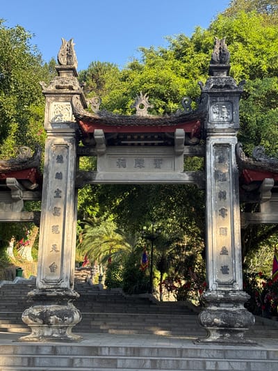 The image captures an ornate stone gate structure, likely an entrance to a temple or historical site, located in Lao Cai Ward, Vietnam. The gate is constructed of gray stone, adorned with traditional Vietnamese architectural features such as intricate carvings, curved roof elements, and dragon-like figures atop the pillars. Inscriptions in Vietnamese characters are visible on the stone surfaces of the gate. The gate opens to a pathway or staircase leading upwards, surrounded by lush green trees and foliage. The sky above is blue, suggesting a clear day. The overall scene conveys a sense of cultural heritage and serene natural surroundings.