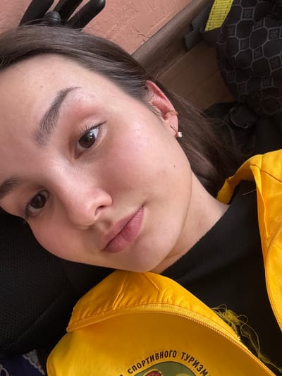 A close-up, angled portrait depicts a female subject, encompassing her face, neck, and upper chest, in an indoor setting. The subject has dark brown hair, dark brown eyes, and fair skin. Her right ear is visible, adorned with a small hoop earring near the helix and a single-stone stud earring on the lobe. Her lips are closed, exhibiting a pink hue. She wears a black garment beneath a bright yellow zippered outer garment, visible in the lower right quadrant. The yellow garment features partially visible embroidered Cyrillic text, reading "СПОРТИВНОГО ТУРИЗМ" (SPORT TOURISM). Loose yellow threads are apparent near the zipper of the yellow garment. The background consists of an out-of-focus reddish-brown textured surface on the upper left and a dark object with a geometric pattern and a yellow strap on the upper right, consistent with a backpack or similar item. The subject maintains direct eye contact with the camera. The image itself contains no discernible features to confirm the specific location of Tayshet, Russia.