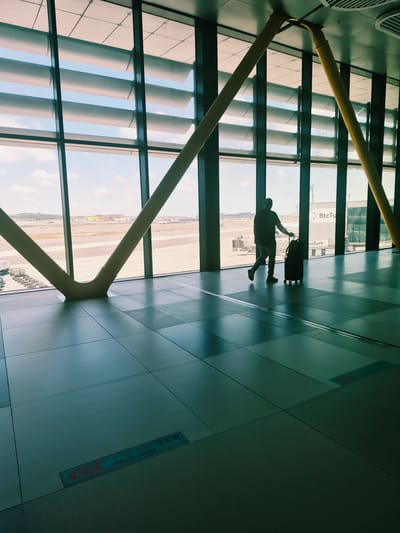 In the city of Arnavutköy, Turkey, a solitary figure walks through a modern airport terminal, their silhouette cast against a large window overlooking an airfield. The individual, clad in a dark t-shirt and jeans, is pulling a dark rolling suitcase. The terminal's architecture is characterized by large, expansive windows with horizontal louvers that filter the daylight, and prominent yellow structural beams that support the ceiling. The floor is tiled in a grid pattern, with subtle directional markers visible. Outside, an airport tarmac is visible, with several aircraft and airport support vehicles. The scene suggests a moment of travel, with the person likely heading to a departure gate or arriving from a flight.