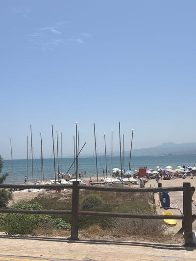 The image shows a view of a beach in Cap-Blanc, Spain on a sunny day. In the foreground, a rustic wooden fence runs horizontally across the frame, separating a paved walkway from a sandy area with dry grass and bushes. Beyond the fence, the beach is populated with many people. Numerous colorful umbrellas provide shade for beachgoers, and several small sailboats are lined up on the sand. The sea is calm and blue, stretching out to the horizon where a hazy coastline with mountains is visible. The sky is clear and pale blue with a few wispy clouds. Several tall, thin masts of sailboats are prominent in the middle ground. Two blue trash receptacles and a yellow object, possibly a small watercraft or toy, are visible near a wooden walkway on the right side of the image. People are seen walking, sitting under umbrellas, and wading in the water.