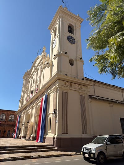 This is a low-angle, eye-level, full shot of the Metropolitan Cathedral of Asunción, Paraguay, on a sunny day with a clear blue sky. The cathedral is a large, cream-colored building with a prominent bell tower that features a clock. Red, white, and blue fabric drapes hang from the front of the cathedral. In the foreground to the right, a silver SUV is parked on the street. To the left of the cathedral, a brick building with arched windows is visible. The street in front of the cathedral is paved, and a stone walkway with stairs leads to the cathedral entrance. A large tree with green foliage is in the upper right portion of the image. There are no people visible in the scene.