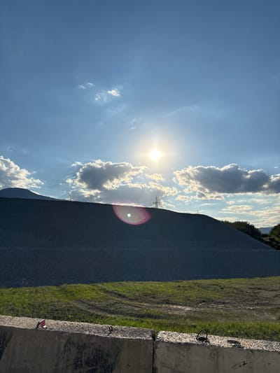 This is a vertical, medium shot photograph taken outdoors. The scene is dominated by a large, dark gray gravel pile that slopes upwards and fills the middle and lower left portions of the frame. The gravel pile is covered in a texture of small, uniform stones. Above the gravel pile, the sky is a clear blue with scattered white clouds. The sun is bright and positioned in the upper center of the frame, casting a lens flare with a pinkish halo and a bright white circle at its center. A single pylon, likely a power line tower, is visible on the crest of the gravel pile, with another smaller structure further to the right. In the foreground, a concrete barrier, possibly a Jersey barrier, runs horizontally across the bottom of the image. The barrier is textured and shows signs of wear, with exposed rebar visible on the right side. In front of the gravel pile, a strip of green grass with some patchy soil is visible. A small red object, possibly a flag or marker, is placed on the concrete barrier in the lower left foreground. The overall scene suggests a construction or industrial site under a bright, sunny sky, possibly in the late afternoon given the direction of the light. The location is not specified beyond "Unknown, Ukraine."