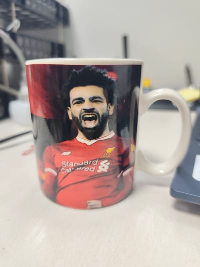 A white ceramic mug with a handle on the right side stands upright on a light-colored, flat surface. The mug features a color print of a male individual identified as Mohamed Salah, depicted from the chest up. He has dark, curly hair and a beard, and his mouth is wide open in a shouting expression, revealing his teeth. He is wearing a red short-sleeved football jersey with white trim, displaying the "Standard Chartered" sponsor logo, an "NB" (New Balance) logo, and the Liverpool FC crest. The background of the print is dark with hints of red. The overall background behind the mug is blurred, showing dark, multi-tiered shelving to the left and a dark electronic device, possibly a keyboard, partially visible to the right. The scene appears to be indoors, likely on a desk. This image was captured in Kangli, Uzbekistan.