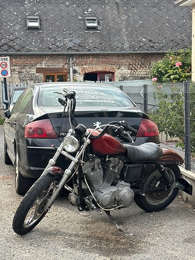 In Arques-la-Bataille, France, a custom-built motorcycle is parked leaning on its kickstand in front of a black Peugeot sedan. The motorcycle has a dark red fuel tank, a black seat, and chrome accents. The sedan is a four-door model with tinted windows. Behind the vehicles, a weathered brick building with a slate roof and two small dormer windows is visible. A metal fence separates the property from a garden with blooming pink roses. A traffic sign indicating a 30 km/h speed limit is posted near the building. The scene is set on a paved street, and the lighting suggests it is daytime.