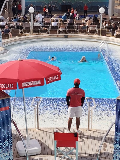 The image depicts a swimming pool scene, likely on a cruise ship, given the layout and design. The pool is rectangular with a blue tiled border. Two people are swimming in the pool. A lifeguard, wearing a red shirt with "POOL SAFETY GUARD" printed on the back, white shorts, and a red cap, stands facing the pool, presumably on duty. A red lifeguard chair is nearby. To the left of the lifeguard is a red umbrella with the word "LIFEGUARD" and "ASSISTENTE BAGNANTI" (Italian for 'lifeguard'). In the background, there are lounge chairs with people relaxing, and beyond that, there are what appear to be other passengers, possibly watching a performance or event. The entire scene appears to be on a cruise ship, not in the city of Cagliari, Italy.