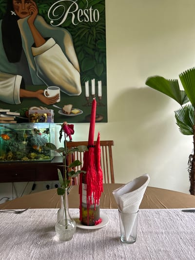 In a well-lit room, likely a restaurant or dining area in Karangasem, Indonesia, a table is set for a meal. A brown tablecloth covers the table, overlaid with a textured white runner. In the foreground, a single red rose wilts in a clear glass vase. To its right, a glass holds white folded napkins. Further back, a wine bottle with melted red wax drips down its sides stands on a white plate. Behind the table, an aquarium filled with goldfish is visible on a dark wooden cabinet. Above the aquarium, a large painting depicts a woman in green and white attire holding a white mug, with the word "Resto" stylized in script above her. Two lit white candles in brass holders are also depicted in the painting. A tall green plant with fan-shaped leaves is visible on the right side of the image. The wall behind the table is painted a pale green.