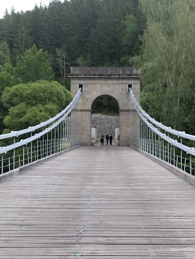 An eye-level, full shot shows a long, wide, wooden suspension bridge stretching towards a stone archway structure with a heavy stone roof. The bridge planks are oriented horizontally and appear weathered, with visible seams and some lighter-colored marks. On either side of the bridge are white suspension cables with vertical supports, attached to the railing which is also white. In the distance, three people are walking away from the camera, toward the archway. They are dressed in casual clothing, with two appearing to be men and one a woman. The archway is built from large stone blocks and has an opening through which the bridge continues. Beyond the bridge and archway, a dense forest of green trees rises up a hillside, with a few lighter green trees in the foreground. The sky is overcast and appears pale. The location is in Dobřejice, Czechia.