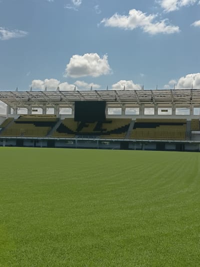 This is a media file showing a wide shot of an empty football stadium from a slightly elevated perspective. The stadium features yellow and black seating, arranged in tiered sections. A large, dark screen is visible above the seating. The stadium structure is modern, with exposed metal supports and roofing. To the right of the stadium, a perfectly manicured green football pitch is visible, showing faint lines from recent mowing. The sky above is a clear blue, dotted with fluffy white clouds, suggesting a daytime setting. The image is taken from a low angle, looking up at the stadium and sky.
The scene is set in Tiraspol City Council, Moldova. No people are present in the stadium, indicating it is either pre-game, post-game, or not in use. The weather appears to be pleasant, with a clear sky and visible clouds, implying it's a fair-weather day. Some text is visible on a banner or sign on one of the stadium's lower levels, reading "НАШ СТАДИОН, НАША КРЕПОСТЬ!". This translates to "Our stadium, our fortress!". The overall impression is one of quiet anticipation and readiness for a sporting event.