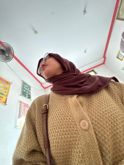 In a brightly lit room, a person wearing a brown hijab and a tan knitted cardigan is captured from a low angle, looking upwards. The person is wearing round-rimmed glasses and has a brown bag strap visible across their chest. The environment appears to be an indoor space with white walls adorned with various posters and a red-trimmed ceiling. A black oscillating fan is mounted on the wall to the left, and a light fixture is visible on the ceiling. The scene suggests a public facility, possibly a health clinic or office, given the informational posters on the wall. The lighting is bright, casting subtle shadows and highlighting the textures of the clothing and the room's surfaces. The overall impression is of a candid moment, perhaps taken while waiting or observing something above. The location is identified as Kebonsari, Indonesia.