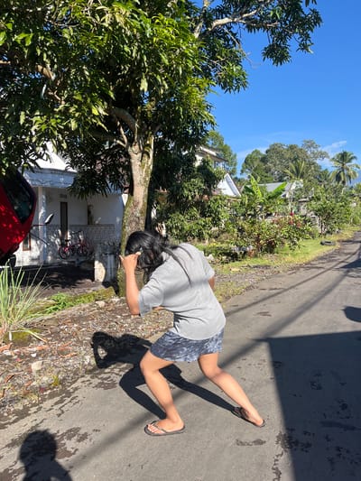 A person with long dark hair, wearing a grey t-shirt and dark patterned shorts, is captured in a low crouch on an asphalt road in Jambearum, Indonesia. Their right arm is raised to their face, obscuring it from view. The person is casting a distinct shadow on the road. To the left of the person, a red vehicle is partially visible, parked near a white house with patterned tiles along its exterior. A pink bicycle is leaning against the wall of the house, and various plants and debris are scattered on the ground. Behind the house, a lush green environment with trees, houses with metal roofs, and palm trees is evident under a clear blue sky. The road extends into the distance, bordered by vegetation and more houses.