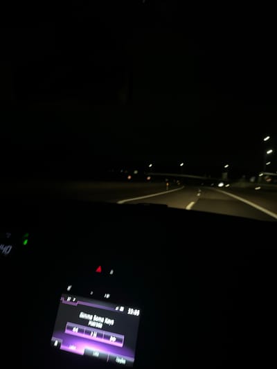 The image captures a nighttime scene from inside a vehicle, likely in Macedo de Cavaleiros, Portugal, presented with a tilted orientation. The setting is primarily dark, illuminated only by artificial lights.
The left side of the frame shows the view through the windshield. Streaks and dots of bright white and warm-toned lights, characteristic of vehicle headlights, taillights, or streetlights, are visible against the dark background, suggesting movement. A faint, curving white line on the ground indicates a road or lane marking.
On the right, a section of the car's interior dashboard and infotainment system is visible. The infotainment screen glows with a purple backlight, displaying media information. Visible text on this screen includes the song title "Gimme Some Keys" and the artist "Maurico." A digital clock on the screen shows the time as "23:05" (11:05 PM). Below the screen, several illuminated controls and indicators are present, including a red triangular light, possibly the hazard light indicator, and a small green light. Partially visible at the very bottom right is the number "140," which may denote the vehicle's speed, potentially 140 km/h.
No people are visible in the image. The scene conveys the activity of driving at night, with music playing, creating an atmosphere of a nocturnal journey.