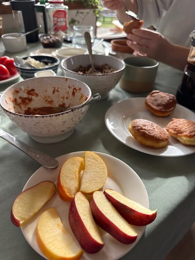 The image depicts an indoor meal setting on a light green tablecloth in Sighnaghi, Georgia. In the foreground, a white plate holds multiple slices of red and yellow apples. To its right, an oval white plate contains three golden-brown, oval-shaped fried pastries. A textured white bowl in the center foreground contains reddish remnants of a dish, with a silver spoon resting inside. Behind it, various bowls are arranged, including one with white crumbly cheese, one with whole red tomatoes, another with dark preserves, and a small white bowl of sugar. A bowl of cereal or granola is also visible. In the upper right quadrant, a person's hands are actively using a spoon to interact with one of the pastries. Other objects on the table include a dark bottle, a two-tone green and beige ceramic mug, and a clear glass of water. Natural light illuminates the scene from the right, casting soft shadows.