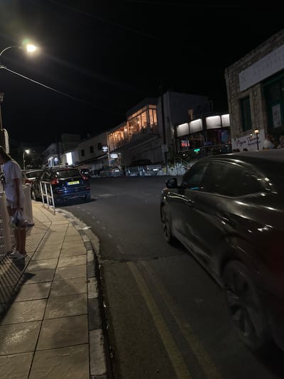 A nighttime street scene in Peyia, Cyprus. On the left, a person in a white t-shirt and white shorts stands on a tiled sidewalk, holding a clear plastic bag with items inside. They are near a white railing. The sidewalk has patterned tiles and a curb. The street is asphalt with yellow double lines indicating a no-passing zone. Several cars are parked on the street and the sidewalk. One dark blue SUV with a license plate reading "LAG 735" is parked on the street. A dark-colored car is in motion in the foreground on the right. Buildings line the street, some with illuminated windows and outdoor seating areas. One building has a sign that reads "Peyia Tavern Special Meze." Another building has a sign that says "PROPERTY VALUERS & CONSULT". A street lamp illuminates the scene from the upper left. The sky is dark.