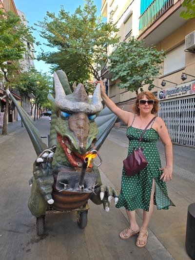 A woman in a green polka dot dress and sunglasses stands next to a dragon float. The float is green and has large horns and blue eyes. There is a flame coming out of the dragon's mouth. The woman is holding onto the dragon's head. The scene is outdoors on a street with buildings and trees in the background. The time of day appears to be daytime, and the weather seems to be clear and sunny. There is text visible on a building in the background that reads "Reparació Calcat Artesania en Cuir". The woman appears to be smiling and enjoying herself. The overall impression is a festive and playful scene, possibly at a street festival or event.