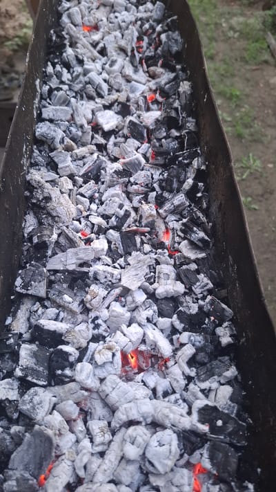 The image shows a close-up, overhead view of glowing charcoal embers in a metal brazier. The embers are a mix of black charcoal chunks and white ash, with visible red-hot areas indicating active burning. The brazier appears to be placed outdoors, with a blurred background suggesting dirt and some vegetation, likely in the city of Shubarșu, Kazakhstan. There are no people or specific actions depicted, only the still life of the burning coals.