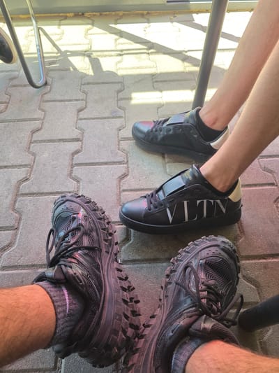 The image shows two pairs of shoes on a paved surface. On the left, a pair of black Valentino sneakers is visible, while on the right, a pair of black Balenciaga sneakers is present. One person is wearing the Valentino sneakers, and another is wearing the Balenciaga sneakers. The paved surface appears to be composed of interlocking bricks or tiles. There are shadows present, which suggests that the photograph was taken outdoors during daylight hours. The location of the image is unknown.